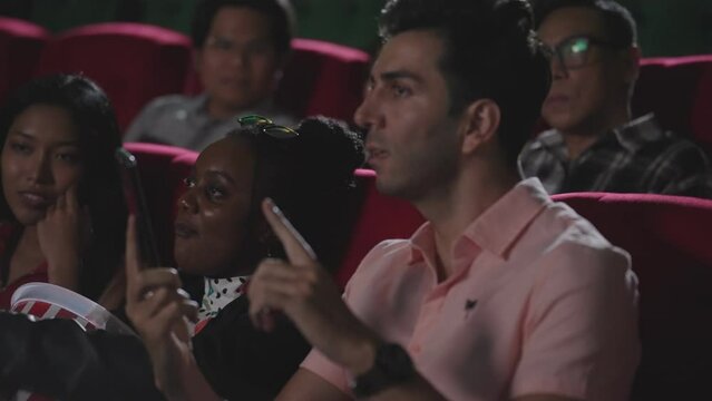 Young hispanic latin man talking on smartphone while watching movie at the cinema and disturbing audience. Phone talks prohibited during movie show. A man disrespectful talking on phone at theater