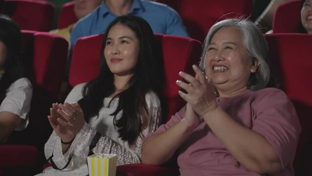 Happy Asian mother and daughter laughing and applauding while watching comedy movie in theater. Group of happy people applauding while sitting on seats in cinema hall enjoying watching movie