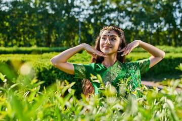 Naklejka na ściany i meble carefree young indian woman in green sari posing near bushes in park in summer