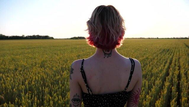 Young female hipster with pink hair standing on green barley field at sunset. Carefree punk girl with tattoos resting at wheat meadow and enjoying freedom. Beautiful scenic landscape at background