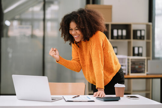 Excited African, American Woman Sitting At Table Feeling Happy Black Woman Overjoyed Accepting Mail At Laptop In Office.