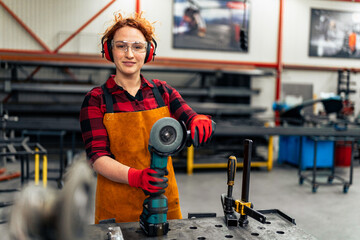 A young girl with curly red hair who is an apprentice in a metal workshop is using tools, she is...