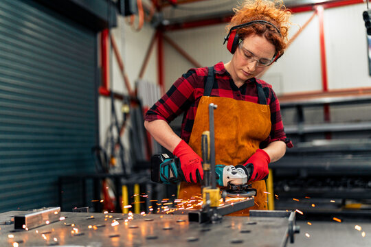 A young girl who is an apprentice in a metal workshop uses a grinder and works on a metal bar, she is wearing protective equipment