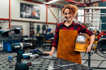 Woman with curly red hair standing in workshop, holding protective equipment under arm and smiling at camera, workshop in background - Powered by Adobe