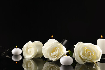 White roses and burning candles on black mirror surface in darkness, space for text. Funeral symbols