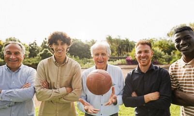 Multi generational men smiling in front of camera - Male multiracial group playing soccer with vintage ball during sunday day at city park - Main focus on center senior man face
