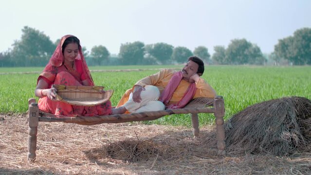 Indian village couple in their field - sitting on a Chaarpai  wheat field  village lifestyle  happy Indian couple. Mid aged Indian desi man wearing kurta pyjama is relaxing and his wife in saree cl...