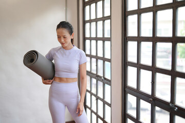 Joyful beautiful Asian woman holding yoga mat with sportswear on a white cement background.