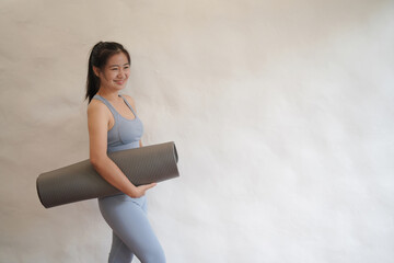 Joyful beautiful Asian woman holding yoga mat with sportswear on a white cement background..