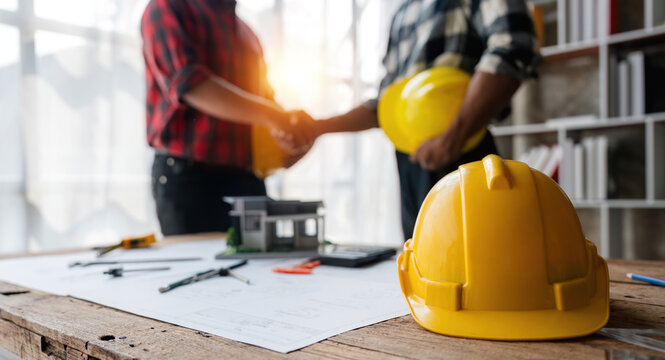 Yellow safety helmet on workplace desk with construction worker team hands shaking greeting start up plan new project contract in office at construction site, partnership and contractor concept
