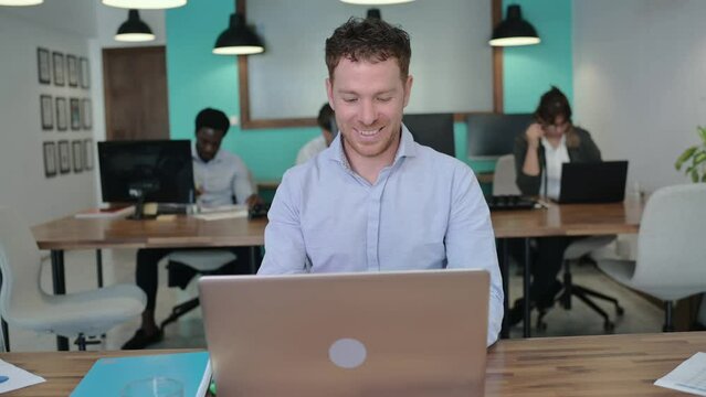Smiling adult businessman in blue shirt typing information on laptop while working in office with colleagues