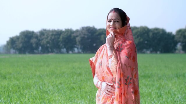 Shot of a married Indian lady standing on her agricultural farm while wearing a traditional printed saree. Beautiful village woman smiling and posing in front of farmland - wheat crops  Indian agri...