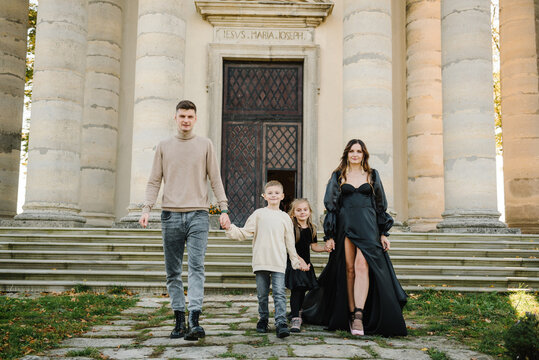 Parents hold hands kids near at Catholic church of Saint Joseph in Pidhirtsi. Mother, father, daughter, son walks on steps near large columns of ancient temple at sunset. Family spending time together