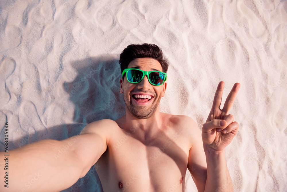 Poster Top view photo of positive funky guy swimwear lying sand showing v-sign recording video vlog outside ocean beach