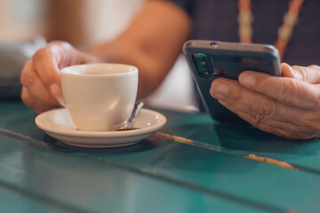 Addicted mature caucasian woman sitting at cafe table with white cup of coffee while using mobile phone. Unrecognizable female enjoying social and technology using smartphone