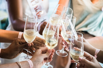 Close-up photo of cheerful multicultural and multiracial girls celebrating a bachelorette party. Bridesmaids' hands clinking champagne glasses indoors.
