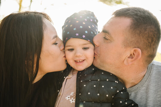 Mom, dad kiss and hug daughter on background of yellow leaves in forest. Upper half. Closeup. Young family spending time together at sunset. Autumn family photo. Happy family walks in autumn park