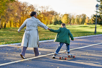 Fototapeta premium boy in outerwear riding penny board and holding hands with mother, african american, autumn leaves