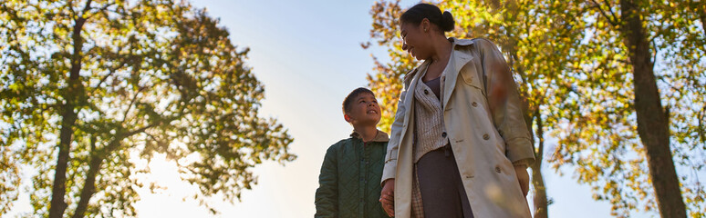 african american mother and child in outerwear holding hands near trees in autumn park, fall, banner