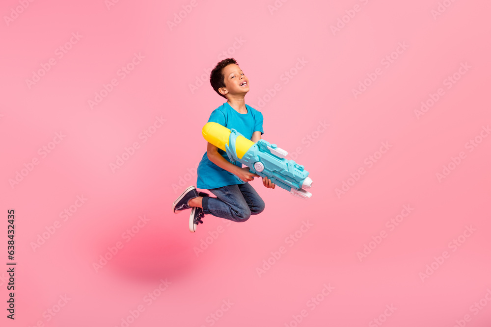 Wall mural Full length photo of cool positive little boy dressed blue t-shirt jumping high shooting water gun isolated pink color background