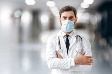 Photo of a caucasian proud male doctor, therapist, surgeon, cardiologist in a medical uniform, protective mask and with stethoscope on his shoulders, standing in a hospital, looking to the camera