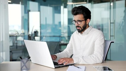 Frustrated young businessman having problems with software or service on laptop computer sitting at workplace in modern office. Worried disappointed worker complains about a bad internet connection
