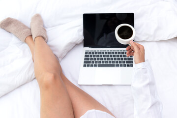 Young people working on laptop and drinking coffee in morning relax mood in winter season. Close up legs women on white bed in the bedroom. Lifestyle Concept.
