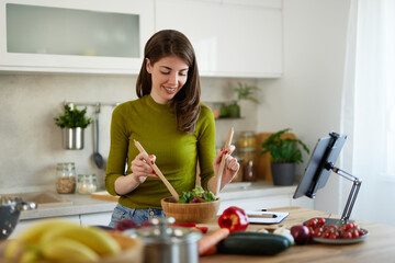 Young woman standing in the kitchen making a salad using online recipe