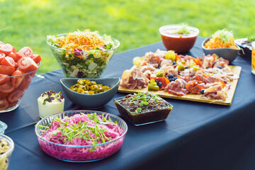 A table is set with delicious snacks prepared for a buffet in the garden. A board with charcuterie, fresh vegetables, vegetable salads, salsa, jalapenos and other treats