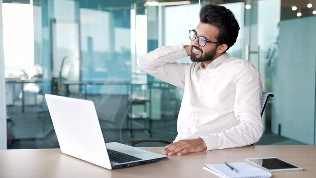 Tired young businessman suffering from neck pain while working on laptop while sitting at desk at workplace in modern office. Sad male employee in a shirt massages and rubs sore muscles, stretches