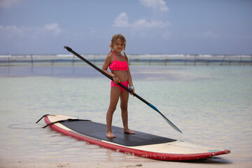Beautiful little girl standing on a paddle board in the ocean at the beach
