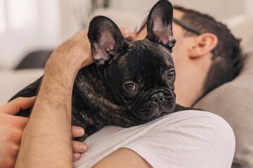 A young positive man is holding a puppy, a black French Bulldog, in an apartment.The concept of...