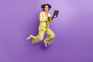 Full length photo of cute shiny woman dressed yellow jacket working modern device jumping high isolated purple color background
