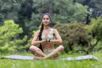  Young Indian woman, yoga instructor and trainer meditating in the park on a mat, sitting in the lotus position in the park with her eyes closed © Tetiana