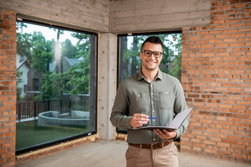 joyful real estate agent in eyeglasses holding folder with pen and looking at camera in new house