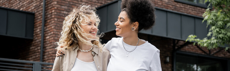 happy and successful interracial lesbian couple looking at each other near own cottage, banner