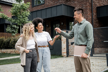 successful realtor with folder giving key from new house to pleased multiethnic lesbian couple