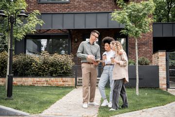 confident real estate broker showing documents to multiethnic lesbian couple near modern cottage