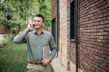 pleased real estate agent with folder looking away while talking on mobile phone near cottage