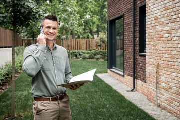 Fototapeta premium cheerful real estate broker with folder talking on smartphone and looking at camera near house