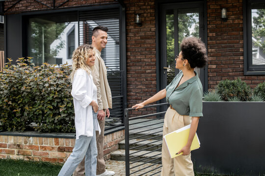 joyful couple talking to african american property realtor near modern building on urban street