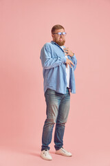Full-length image of bearded man in casual clothes standing with unpleasant meme face against pink studio background. Concept of human emotions, lifestyle, facial expression, ad. Copy space for ad