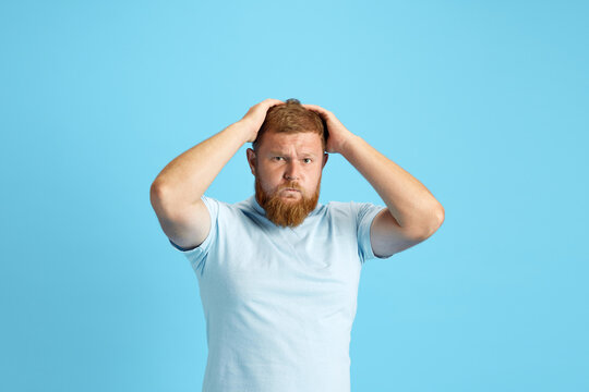 Emotional bearded man holding head with hands standing with shocked and stressful face against blue studio background. Concept of human emotions, lifestyle, facial expression, ad. Copy space for ad