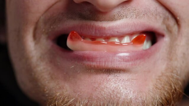A young bearded man holds gummy candy in his mouth shaped like red vampire fangs and eats them.