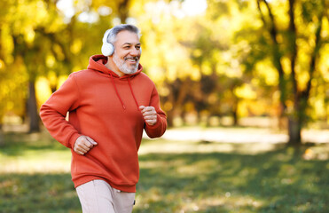 Fototapeta premium Happy senior man in headphones listening music while jogging outside in city park