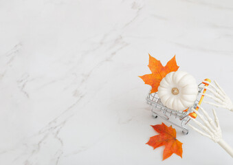 white halloween pumpkin in little grocery trolley with skeleton arms and maple leaves on marble background
