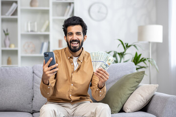 Male winner of online game sitting on sofa at home, hispanic joyfully shouting and smiling looking at camera, holding phone and money cash dollars, using app on smartphone