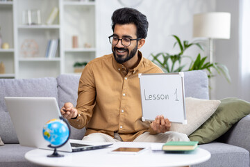 Young male teacher teaching remotely from home, hispanic using laptop for video call and communication and teaching students remotely, explaining new school material smiling