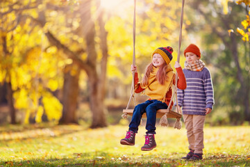 Boy swinging smiling girl on the rope swing in the autumnal park.