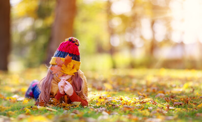 Playful little girl hiding her face behind autumnal leaf in the park.